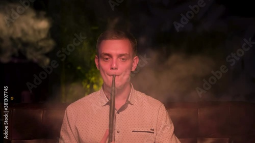 Caucasian man exhaling a fog cloud and chilling. Media. Front view of handsome young guy smoking hookah alone and feeling depressed at dark room of shisha lounge. photo