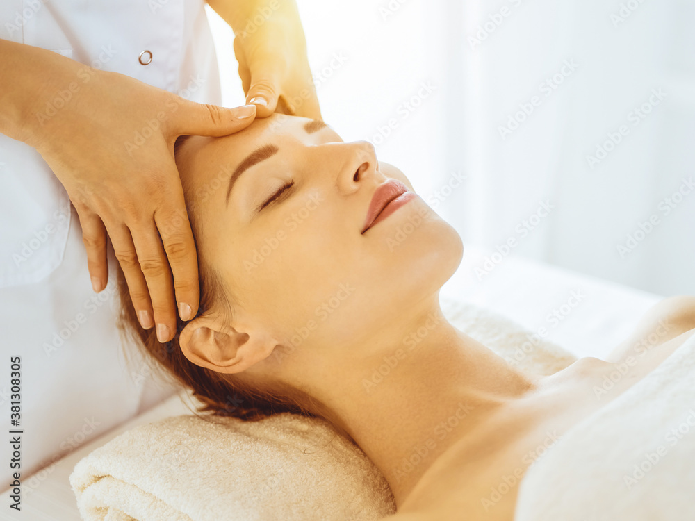 Beautiful woman enjoying facial massage with closed eyes in sunny spa center. Relaxing treatment concept in medicine
