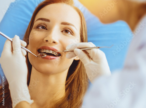 Smiling young woman with orthodontic brackets examined by dentist in sunny dental clinic. Healthy teeth and medicine concept