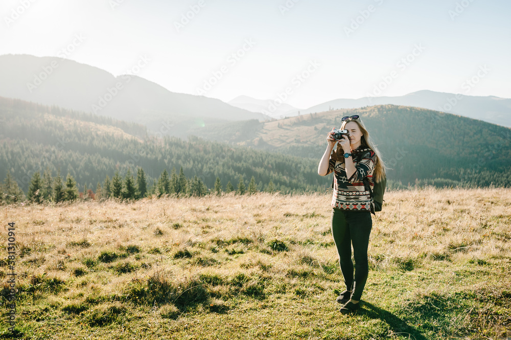 Hipster girl in sunglasses with camera and backpack travels and enjoys nature. Hiker in the mountains at sunset. Woman photographing the sunrise.