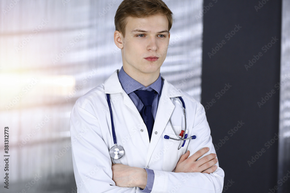 Young doctor standing with crossed arms in sunny clinic near his working place. Medicine concept Young doctor standing with crossed arms in sunny clinic near his working place. Medicine concept