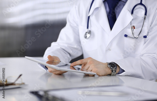 Doctor sitting and working with tablet computer in clinic at his working place, close-up. Young physician at work. Perfect medical service, medicine concept