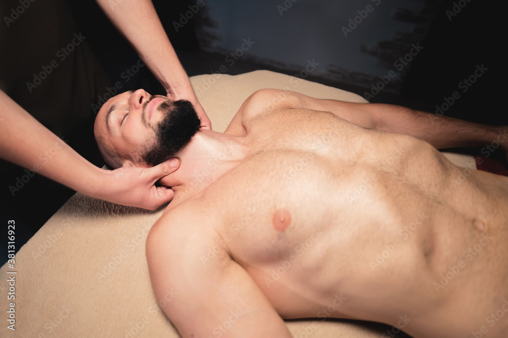 Masseur giving neck massage to young wealthy caucasian athlete in dark room of spa salon Masseur giving neck massage to young wealthy caucasian athlete in dark room of spa salon
