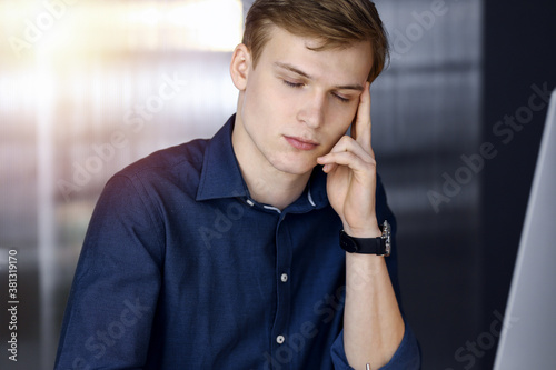 Young blond businessman feeling stress at workplace in a darkened office  glare of light on the background. Startup business means working hard and out of time