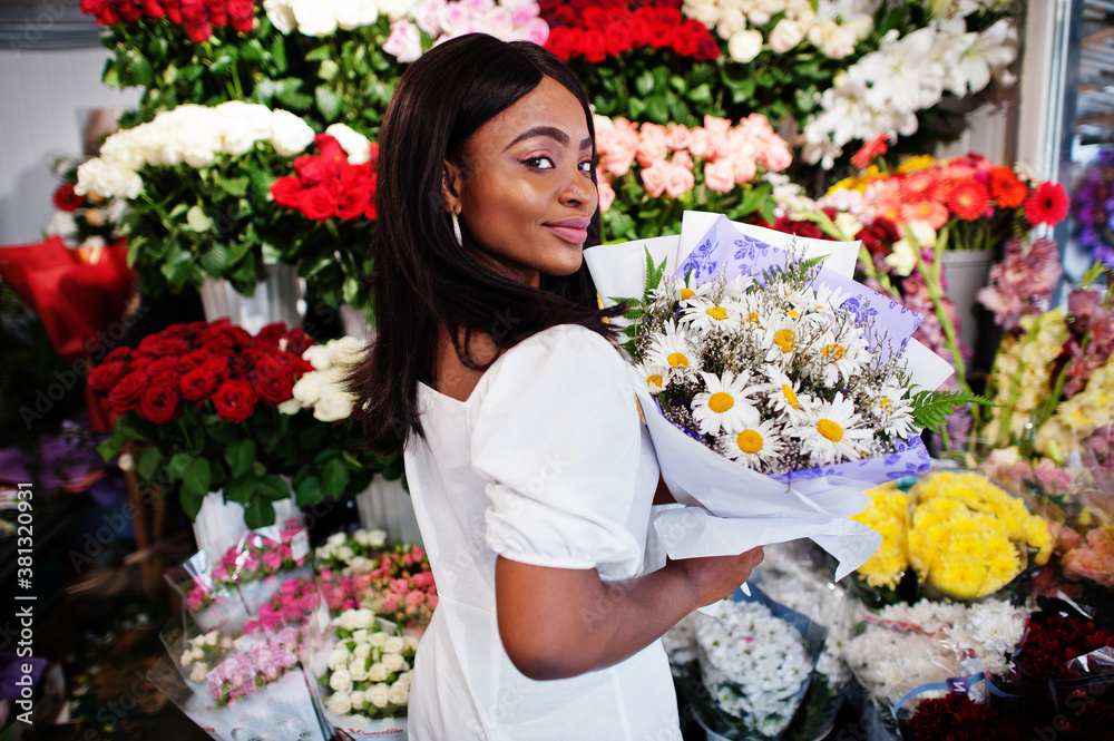 Beautiful african american girl in tender white dress with bouquet flowers in hands standing against floral background in flower shop.Black female florist.