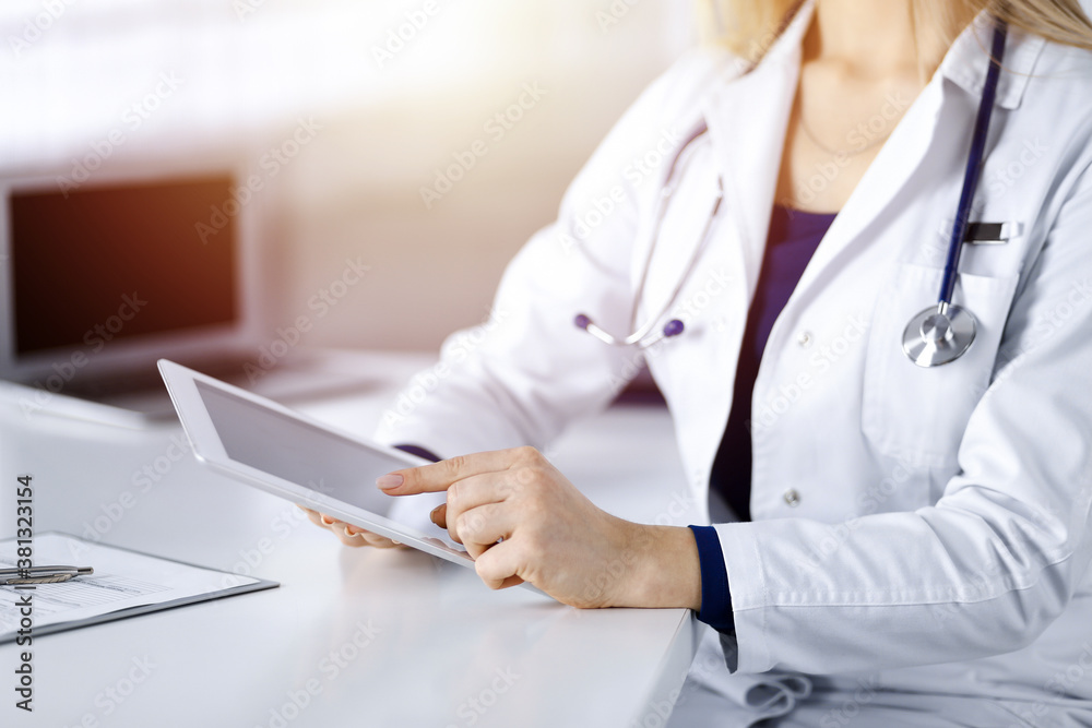 Unknown young woman-doctor is checking some medication names, while sitting at the desk in her sunny cabinet in a clinic. Physician with a stethoscope is using a tablet computer, close-up. Perfect Unknown young woman-doctor is checking some medication names, while sitting at the desk in her sunny cabinet in a clinic. Physician with a stethoscope is using a tablet computer, close-up. Perfect