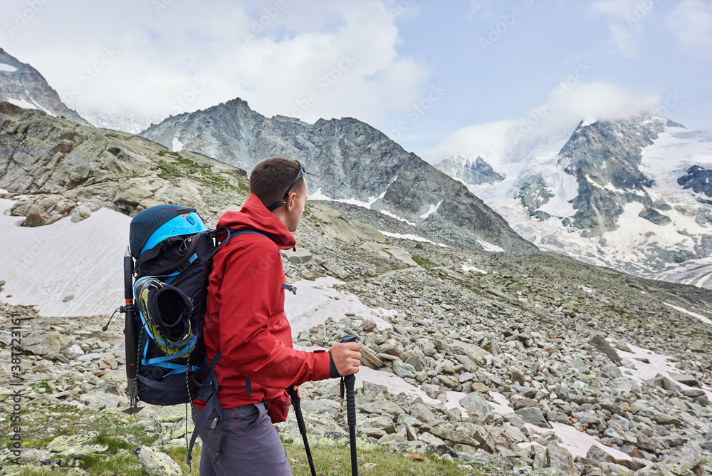 Side view snapshot of a man wearing a red jacket and a backpack, having a walk with hiking poles in the mountains situated in Swiss Alps, enjoying beautiful scenery Side view snapshot of a man wearing a red jacket and a backpack, having a walk with hiking poles in the mountains situated in Swiss Alps, enjoying beautiful scenery