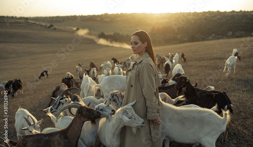 Shepherdess stands in a pasture among goats at sunset.