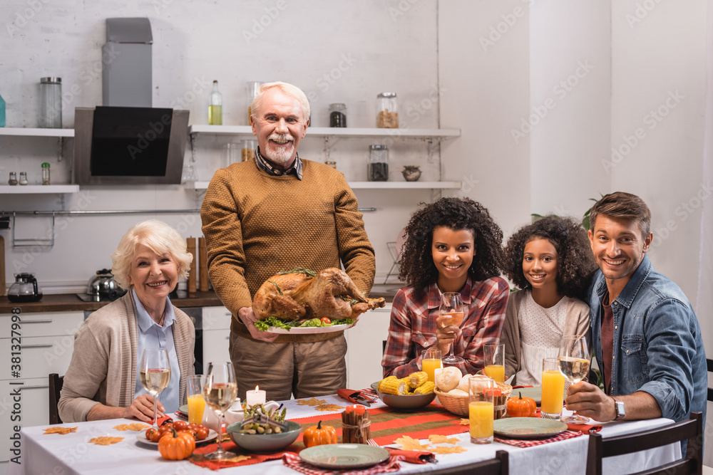 Selective focus of elderly man holding turkey near multiethnic family with wine during thanksgiving