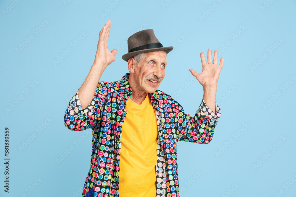 Shocked, astonished. Portrait of senior hipster man in stylish hat isolated on blue studio background. Tech and joyful elderly lifestyle concept. Trendy colors, forever youth. Copyspace for ad. Shocked, astonished. Portrait of senior hipster man in stylish hat isolated on blue studio background. Tech and joyful elderly lifestyle concept. Trendy colors, forever youth. Copyspace for ad.