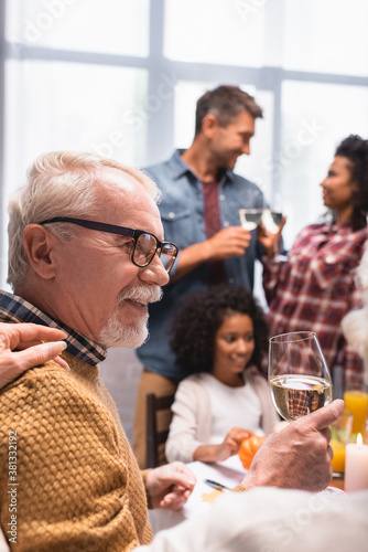 Selective focus of elderly man holding glass of wine while celebrating thanksgiving with multicultural family at home