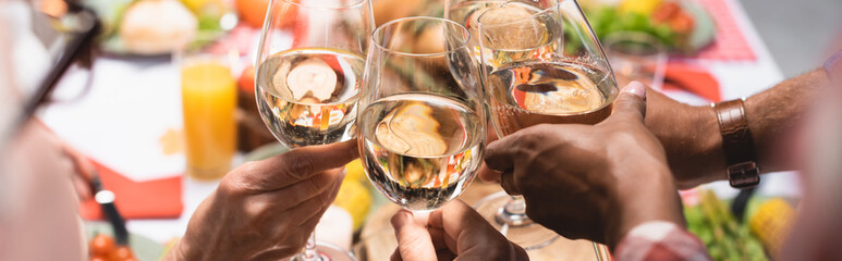 cropped view of multiethnic family clinking glasses of white wine during Thanksgiving dinner, horizontal image