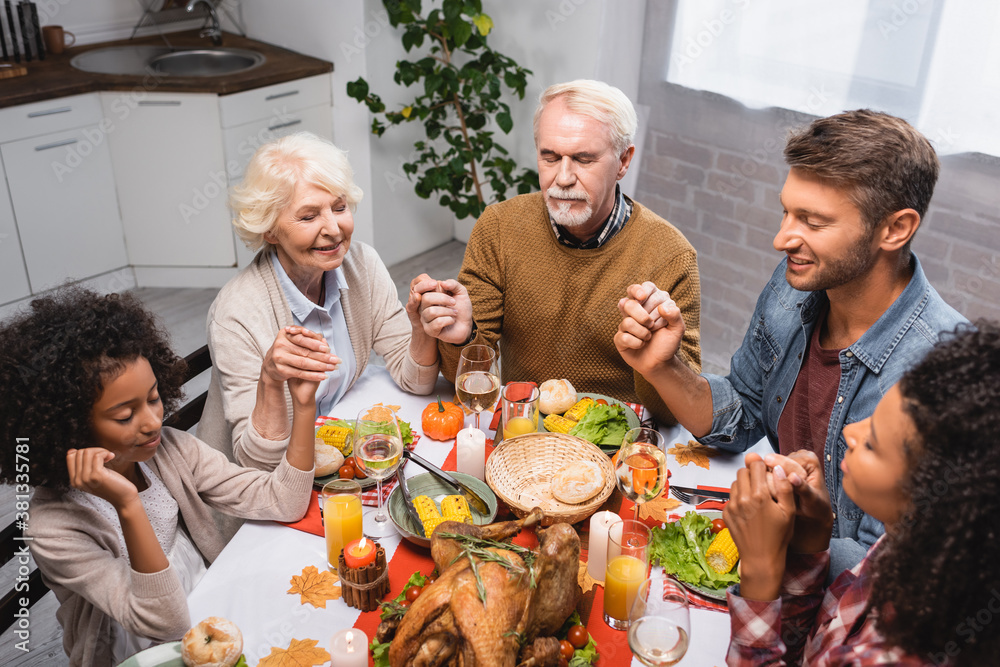 multiethnic family with closed eyes holding hands and celebrating thanksgiving at home