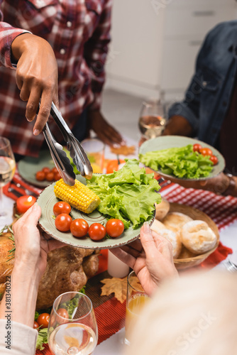 partial view of senior woman holding plate with corn  lettuce and cherry tomatoes during thanksgiving dinner with multiethnic family