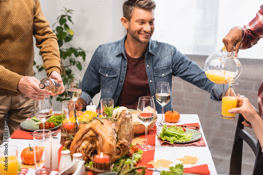 excited man sitting at table served with thanksgiving dinner while multicultural relatives pouring drinks into glasses