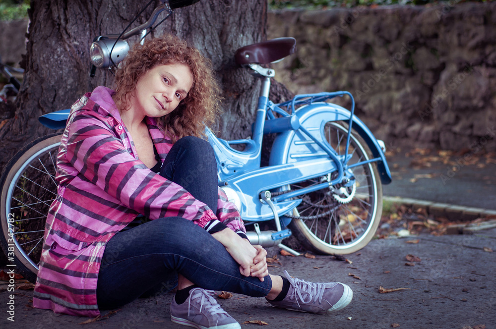 Obraz premium Romantic curly happy young woman sits near her retro bike outdoors under the tree in autumn. People and Transport