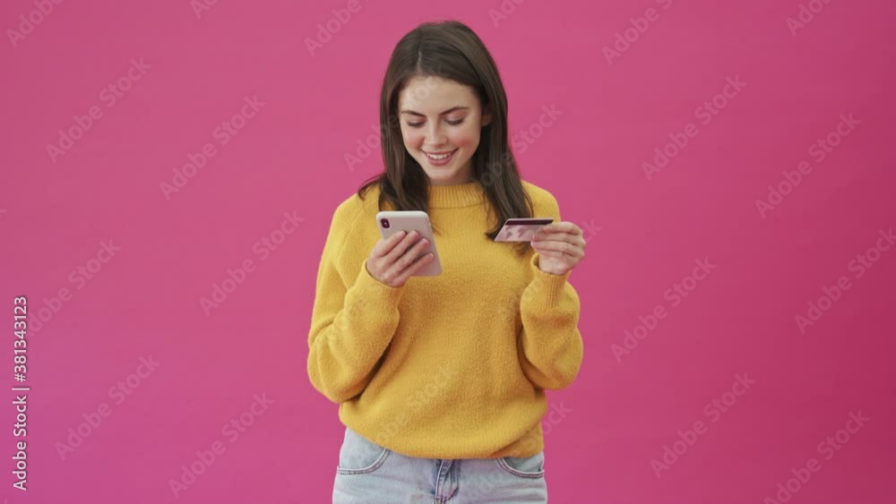 A smiling woman in a yellow sweater is using her smartphone while holding a credit card to transfer money standing isolated over a pink background A smiling woman in a yellow sweater is using her smartphone while holding a credit card to transfer money standing isolated over a pink background