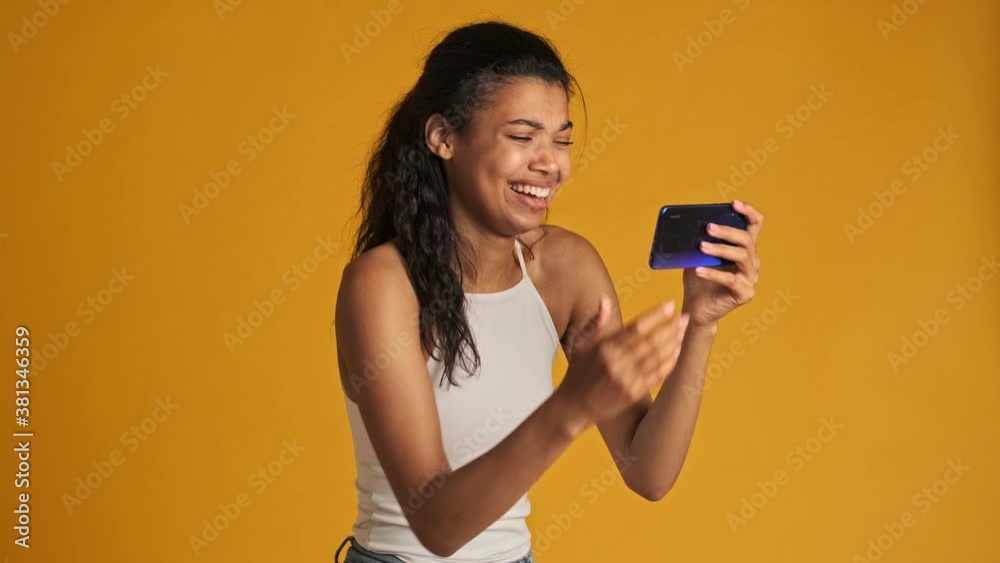 A laughing young woman is watching funny video on her smartphone holding it horizontally standing isolated over yellow background A laughing young woman is watching funny video on her smartphone holding it horizontally standing isolated over yellow background