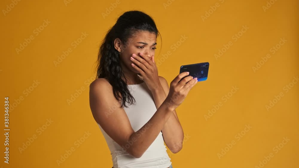 A scared young woman is watching frightened video on her smartphone holding it horizontally standing isolated over yellow background A scared young woman is watching frightened video on her smartphone holding it horizontally standing isolated over yellow background