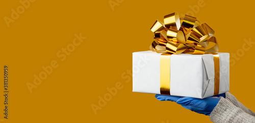 Christmas gift. Pandemic protection. Safety delivery. New Year 2021. Female hands in medicine gloves holding present box taped golden ribbon isolated on yellow copy space. Advertising background.