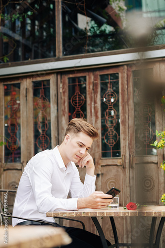 Portrait of pensive young man sitting at table in outdoor cafe, waiting for girlfriend and checking messages in smartphone