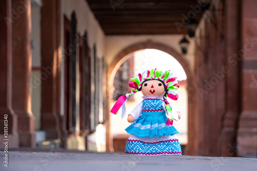 Muñeca de trapo artesanal tradicional con fondo de arco arquitectónico y columnas.