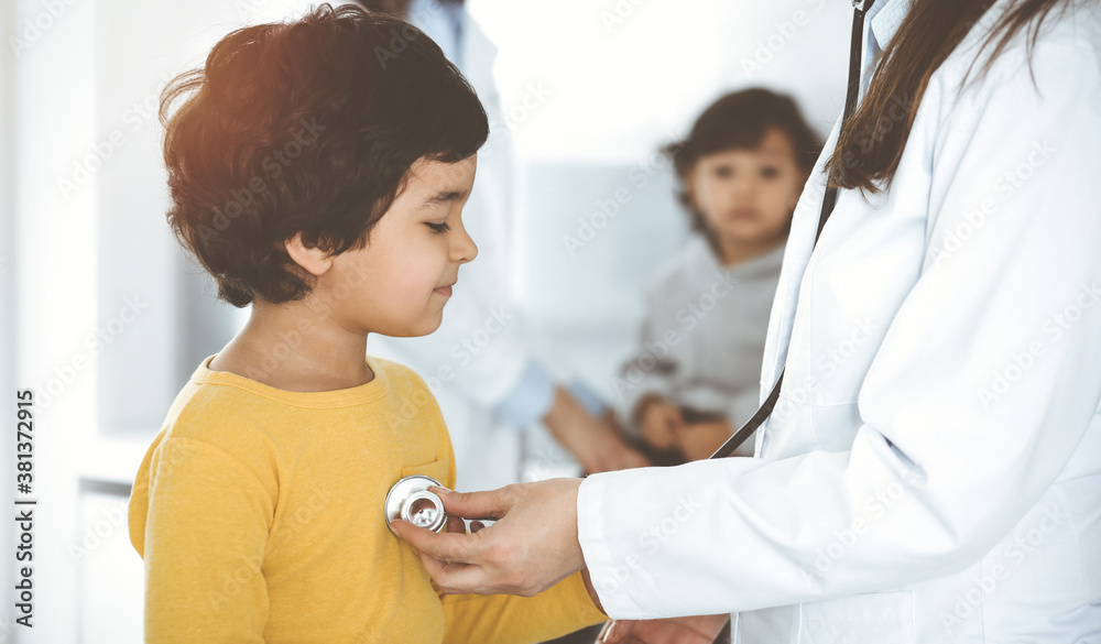 Obraz premium Woman-doctor examining a child patient by stethoscope in sunny clinik. Cute arab boy at physician appointment