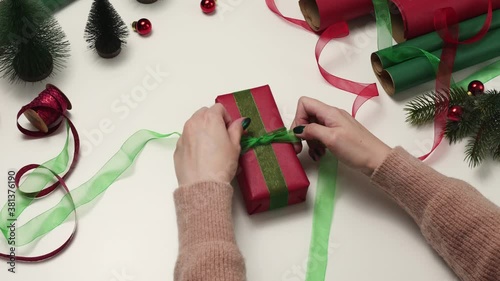 Female hande tying green ribbon on gift wrapped in craftool red paper. Woman packing present on a white table. Preparing for Christmas holidays, photo