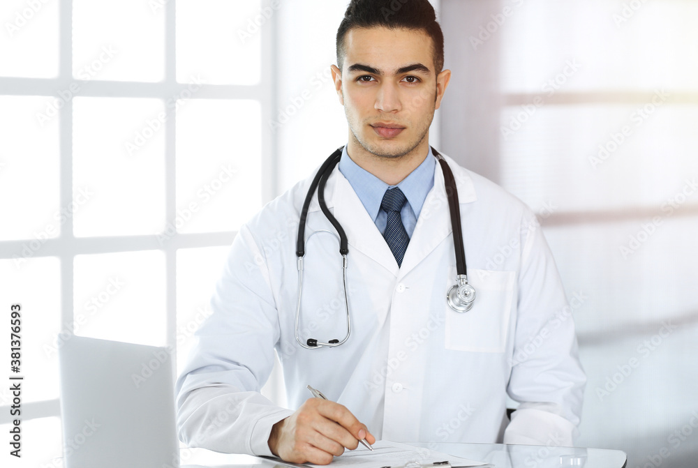 Arab doctor man is using laptop computer while filling up medication history records form sunny clinic. Medicine concept Arab doctor man is using laptop computer while filling up medication history records form sunny clinic. Medicine concept