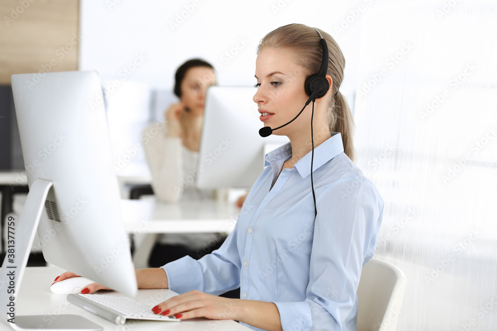 Blond woman call operator is using computer and headset for consulting clients online. Group of diverse people working as customer service occupation. Business concept Blond woman call operator is using computer and headset for consulting clients online. Group of diverse people working as customer service occupation. Business concept