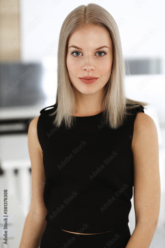 Young blond businesswoman is standing straight in modern office. Business people concept. Black dress do suits to her much at this portrait