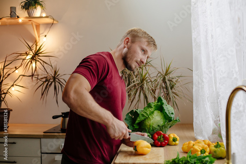 Happy handsome man is cooking in the kitchen by the window. Cut vegetables. Fresh bell peppers, cabbage and mint. Harvesting. selective focus
