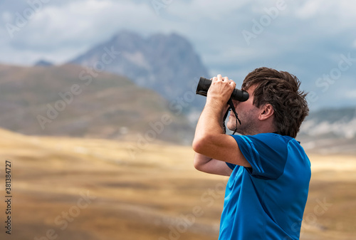 Portrait of a man standing on a mountain hill and looking into the binoculars in the distance, Campo Imperatore, Gran Sasso National Park, Abruzzo region, Italy. Travel concept. 