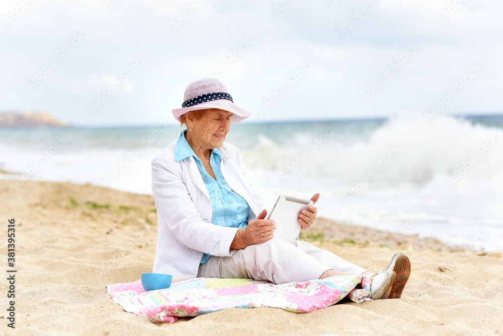 An elderly woman sits on the seashore with a tablet.