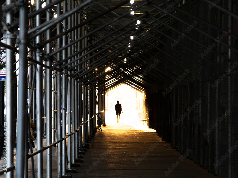 Man walking down the city sidewalk under construction scaffolding with light glowing in the background