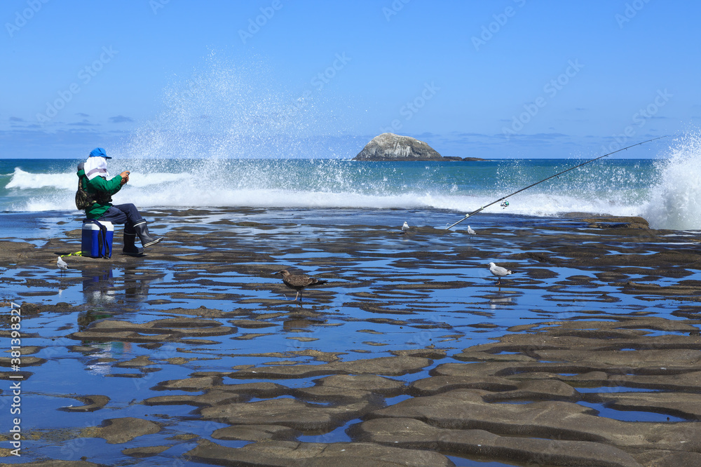 Naklejka premium Man fishing from a rocky shore, with waves breaking behind him. Muriwai Beach, New Zealand