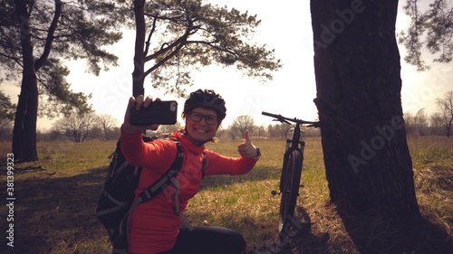 Photos Young caucasian woman athlete tourist cyclist uses hand smart phone photo of herself selfie sitting near tree in coniferous forest outside the city