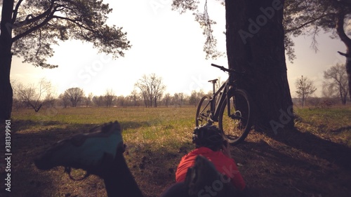 Young caucasian woman athlete tourist cyclist makes photo on phone, photographs his bike standing near tree on nature field sunny day. Girl in helmet and sportswear resting break mountain bike photo
