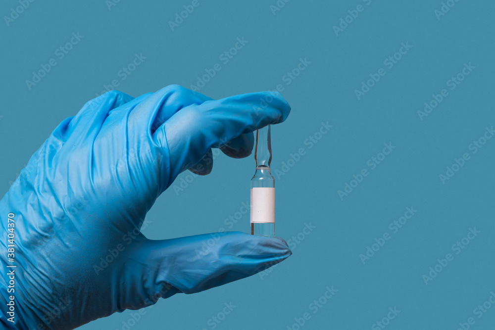 A doctor's hand in a blue glove holds a bottle of COVID-19 medicine or vaccine on a blue background A doctor's hand in a blue glove holds a bottle of COVID-19 medicine or vaccine on a blue background