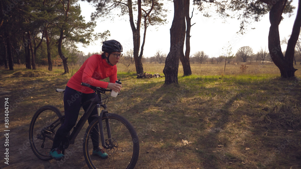 Obraz premium The topic health problems with athletes. Caucasian woman cyclist on mountain bike finish workout in forest. Emotion tired, hard fast heart beating and hard to breathe. Cardiac pathology tachycardia
