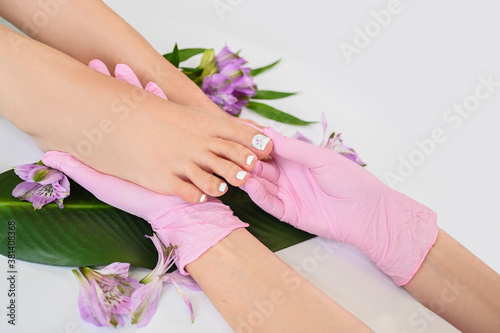 Beautiful perfect skin female legs feet top view with tropical flowers and green palm leaf. Hands of beautician in pink gloves. Nail polish, care and clean, spa pedicure treatment in white. Copy space