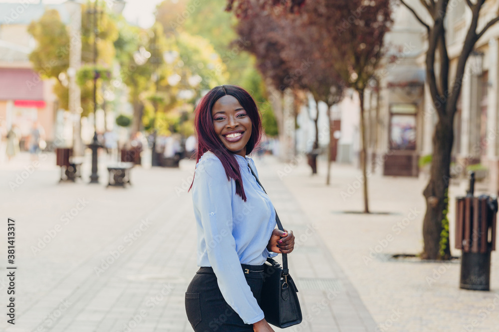 Fototapeta premium a smiling young African American girl with pink hair and a blue shirt walking outside on a sunny day.