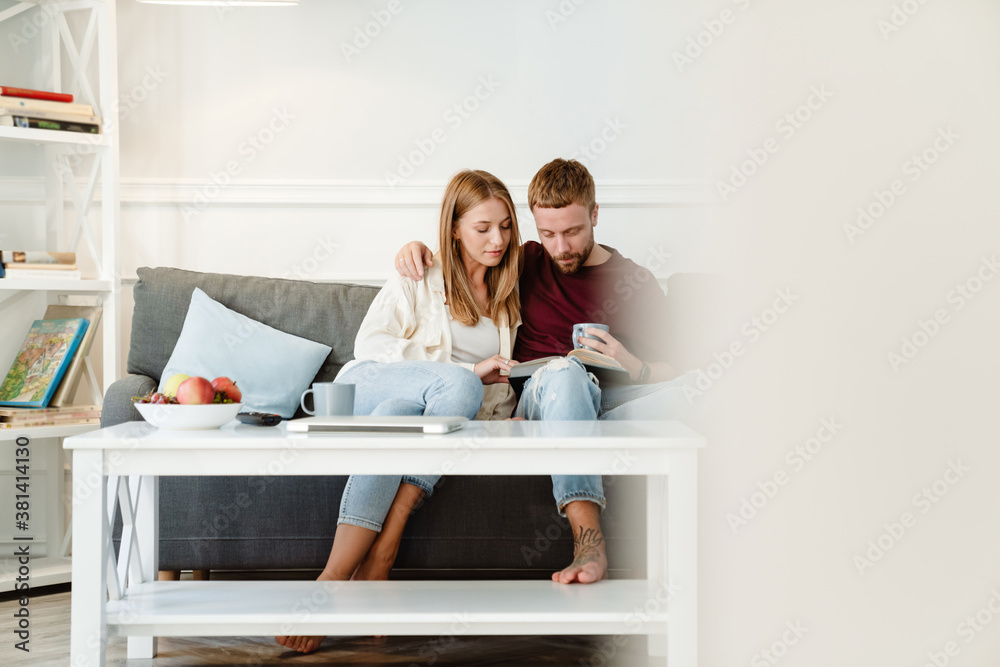 Photo of focused couple hugging and reading book while sitting on couch Photo of focused couple hugging and reading book while sitting on couch