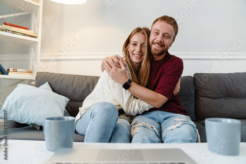 Image of ginger couple hugging and using laptop while sitting on sofa