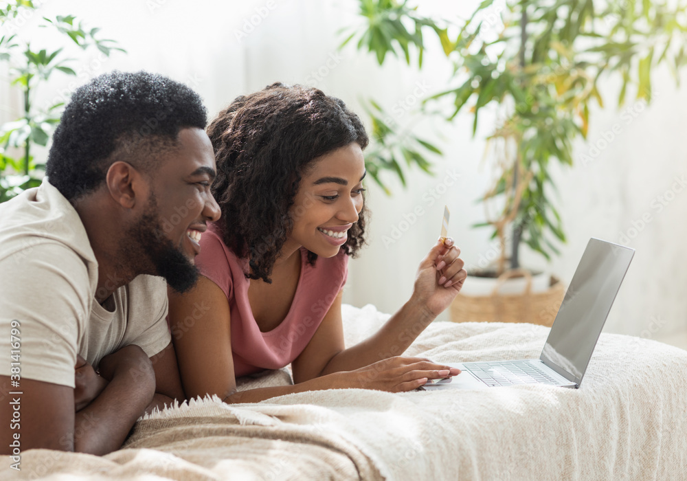 Cheerful black couple using laptop, lying on bed