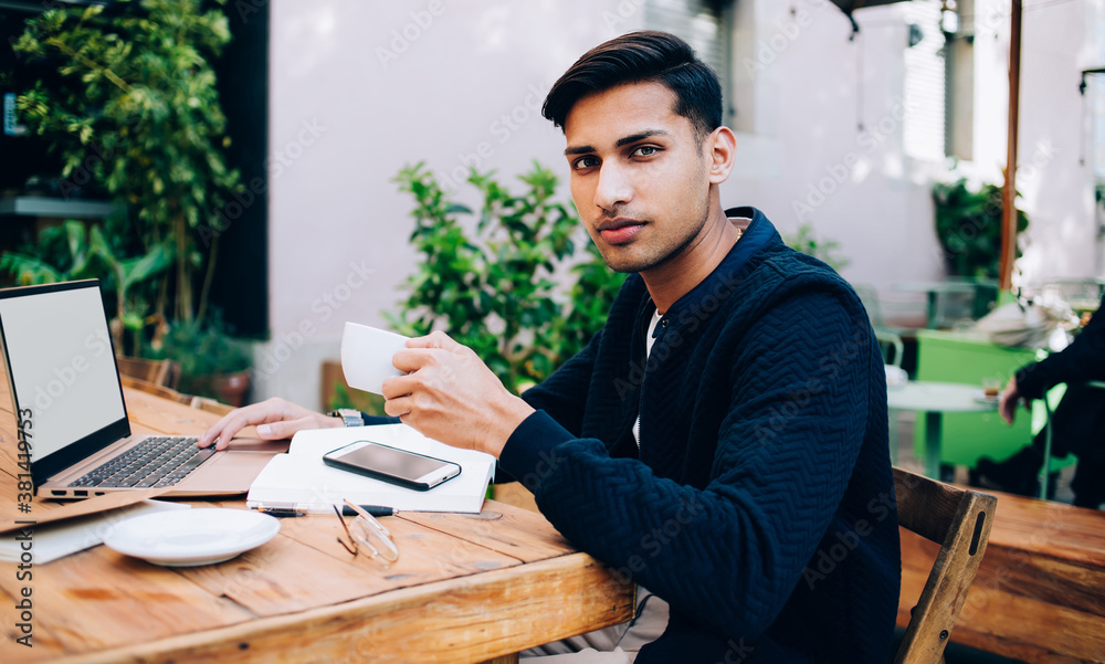 Prosperous hindu male entrepreneur in trendy wear sitting at table with laptop computer creating publications for share in blog, confident male proud ceo drinking coffee on terrace during distance job Prosperous hindu male entrepreneur in trendy wear sitting at table with laptop computer creating publications for share in blog, confident male proud ceo drinking coffee on terrace during distance job
