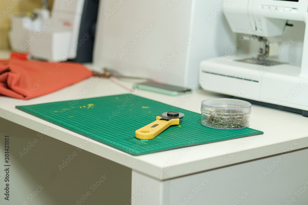 Seamstress's desk - cloth cutting tools - knife, pins and other accessories, a piece of orange cloth and white electric sewing machine in the background in blur Seamstress's desk - cloth cutting tools - knife, pins and other accessories, a piece of orange cloth and white electric sewing machine in the background in blur
