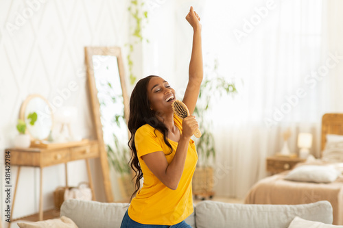 Cheerful black woman with hairbrush as mic singing her favorite song and dancing at home