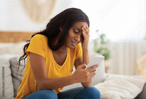 Fotografia Unhappy African American lady looking at her smartphone with disgust indoors, co