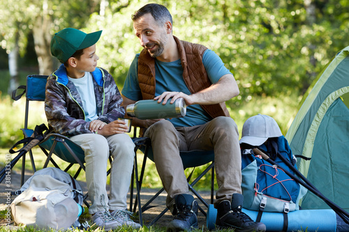 Full length portrait of loving father and son sharing hot drink while enjoying camping together in nature, copy space